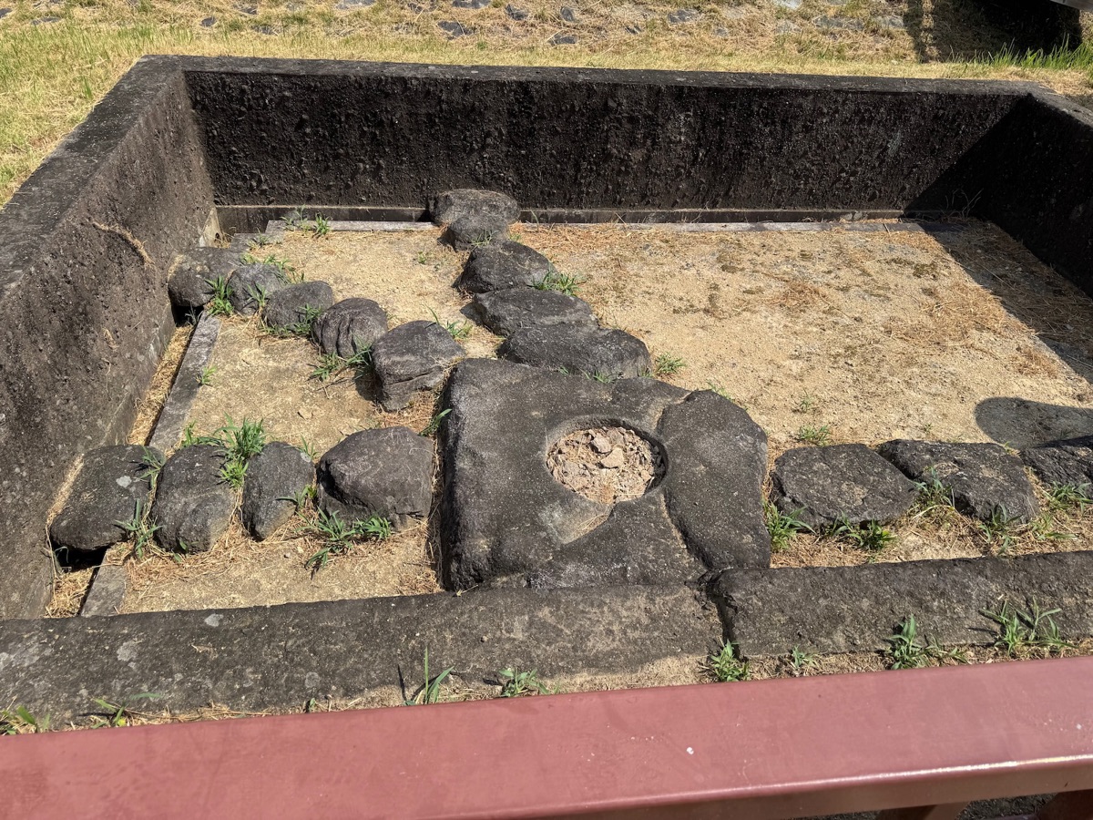 地面を深く掘り下げた底に、石が縦横の列をなして並ぶ。中ほどの平らな四角い石には、その中央に丸い穴があいている。